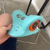 Imagem enviada pelo cliente. Clique para ver mais detalhes. A imagem representa: Um dispositivo eletrônico de massagem nos pés turquesa com furos e um display digital laranja sendo segurado na mão de alguém